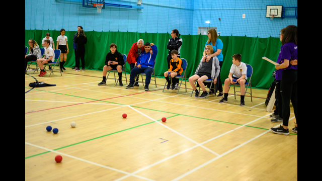 Children sat in chairs in a sports hall playing boccia