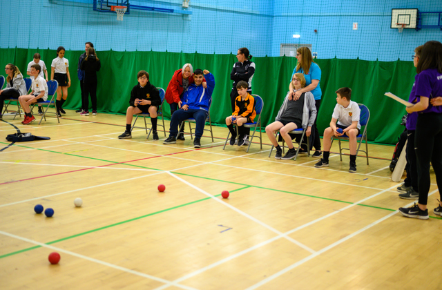 Children sat in chairs in a sports hall playing boccia