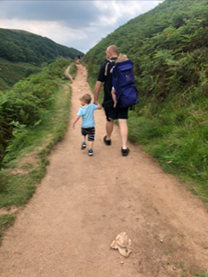 Man walking along a path in hilly green space holding a child's hand with another child in a baby backpack