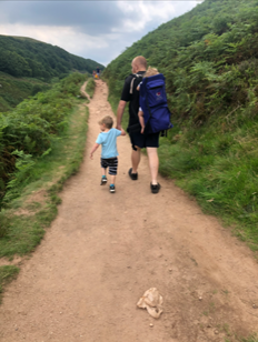 Man walking along a path in hilly green space holding a child's hand with another child in a baby backpack