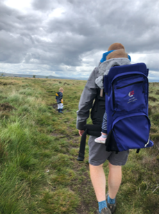 Man walking through a large grassy field with a child in a baby backpack and another slightly further in front of him along the path