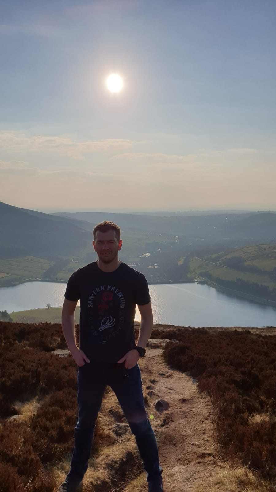 Man stood on a path in green space with a rural panorama behind him