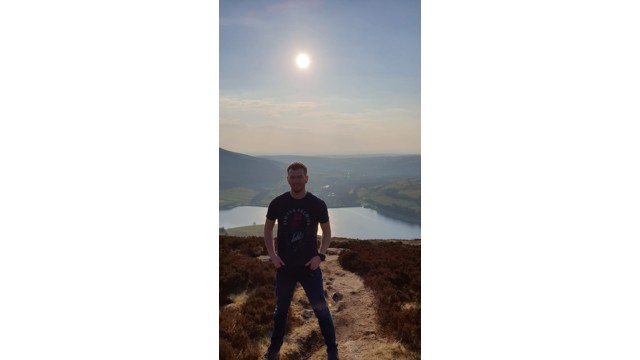 Man stood on a path in green space with a rural panorama behind him